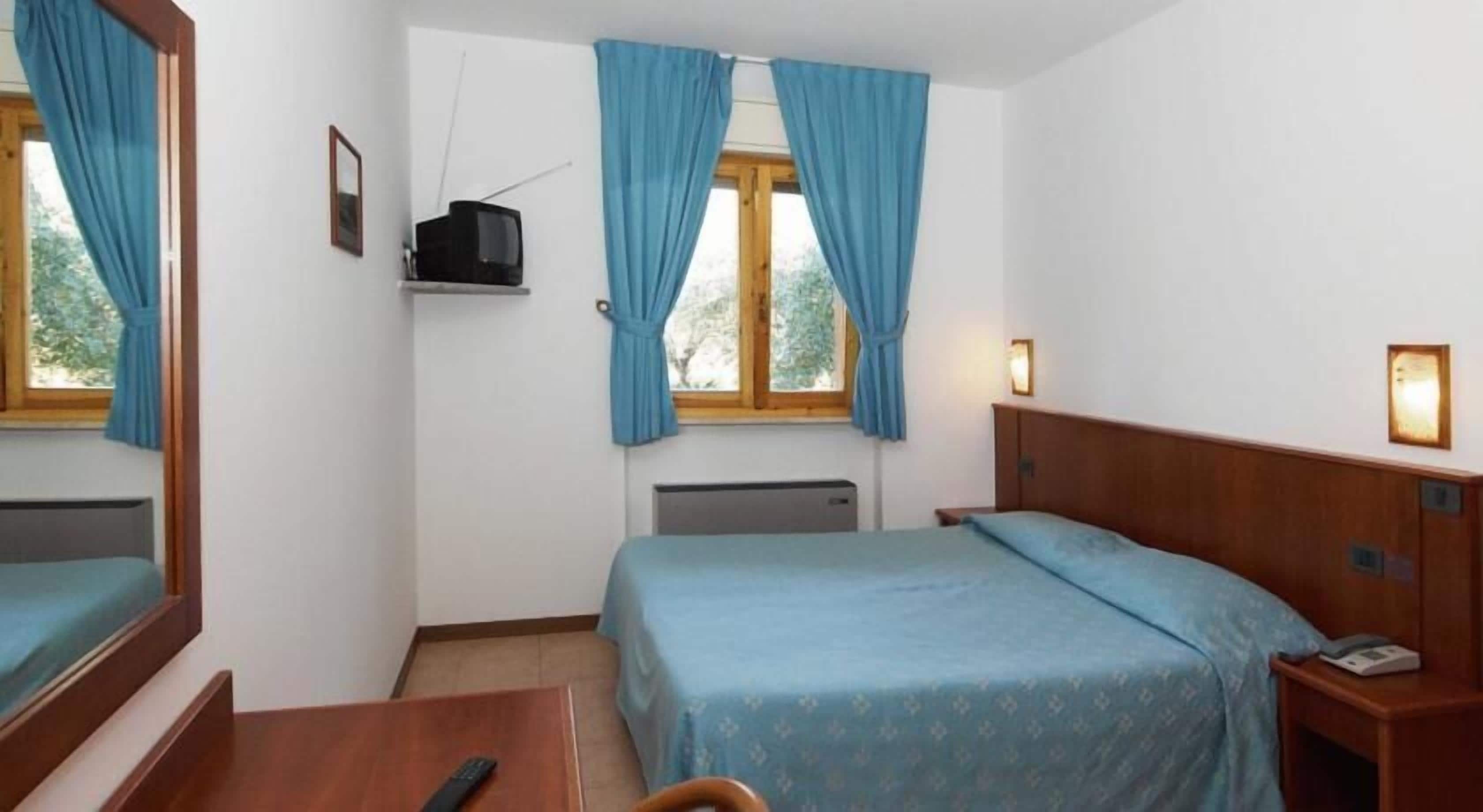 Quarto de hotel com cama, escrivaninha, cadeira, televisão e janela com cortinas.