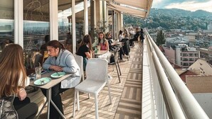 Ein Essbereich im Freien mit Tischen und Stühlen, großen Fenstern und Blick auf die Stadt.