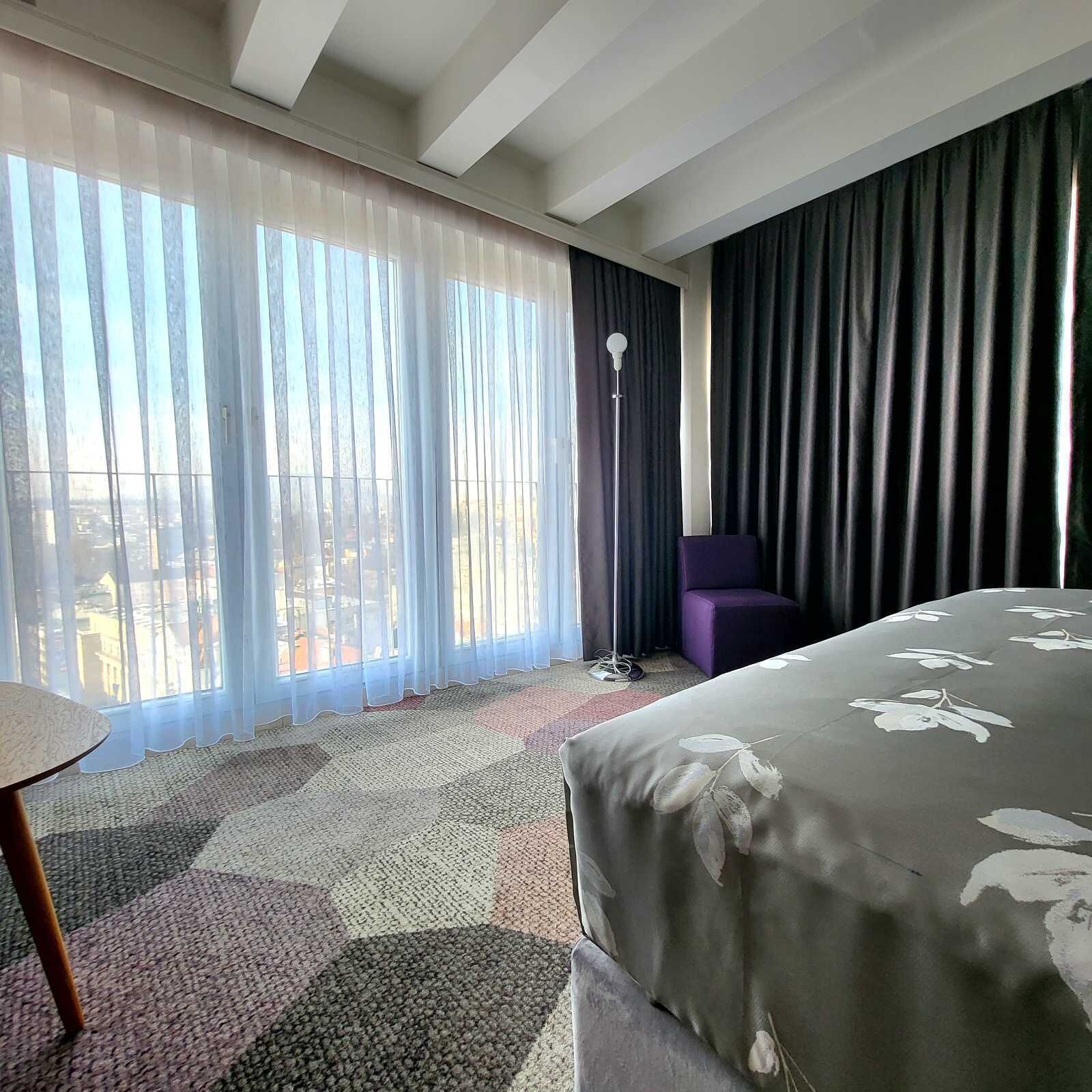 Une chambre d’hôtel avec un lit, une chaise, une lampe sur pied et une vue sur la ville à travers des rideaux transparents.