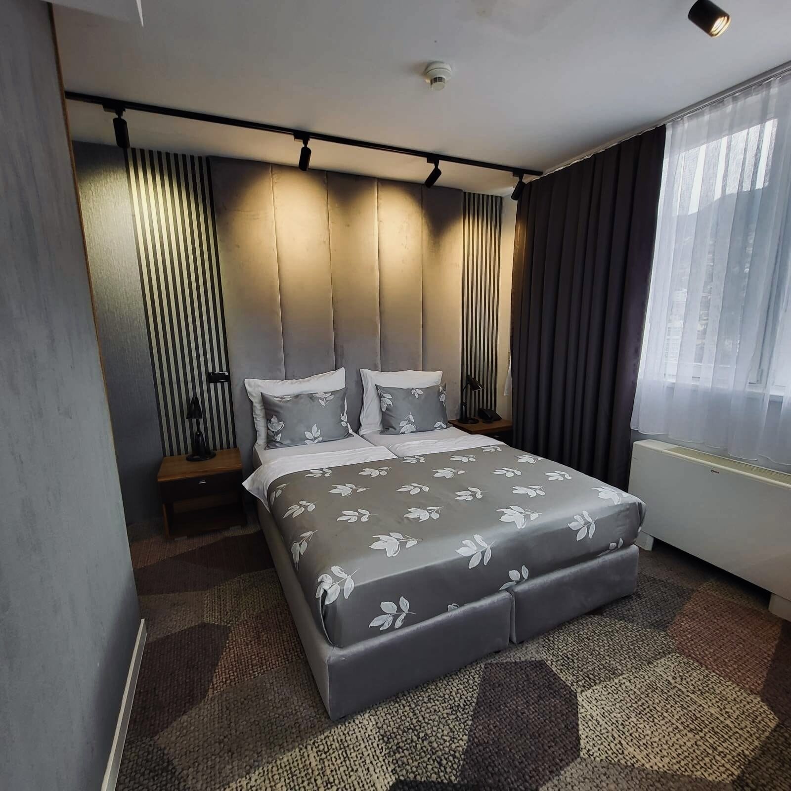 Une chambre d’hôtel moderne avec un grand lit, une table de chevet, une lampe et une fenêtre avec des rideaux.