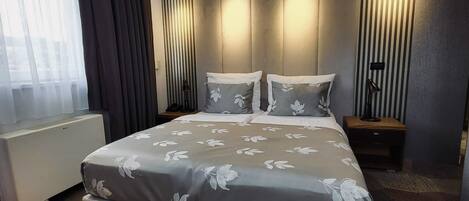 A modern hotel room with a large bed, a bedside table, a lamp, and a view of the outdoors through the window.