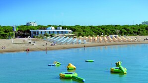A beach with rows of umbrellas and lounge chairs, a building with a blue facade, and inflatable water toys in the water. - Union Lido Art & Park Hotel (Cavallino-Treporti)