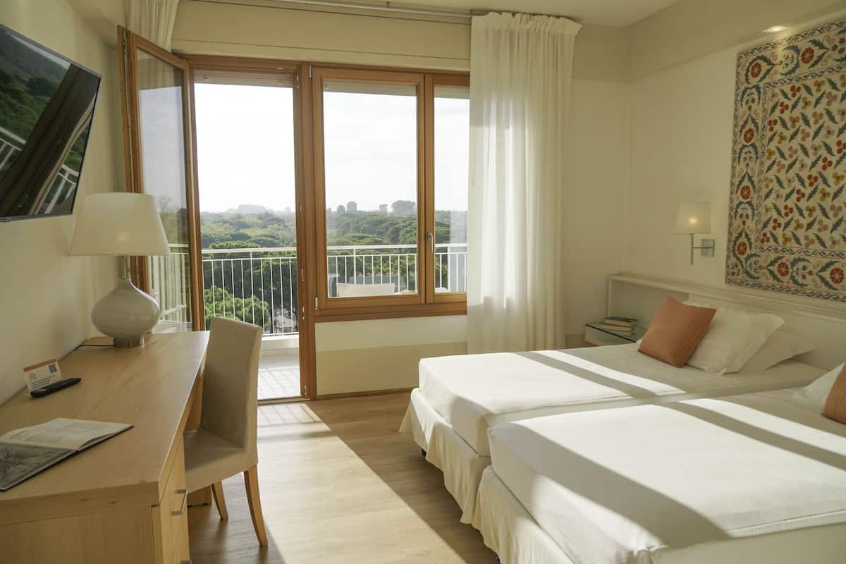 A hotel room with two beds, a desk with a lamp, a chair, a nightstand, and a view of greenery through the window.