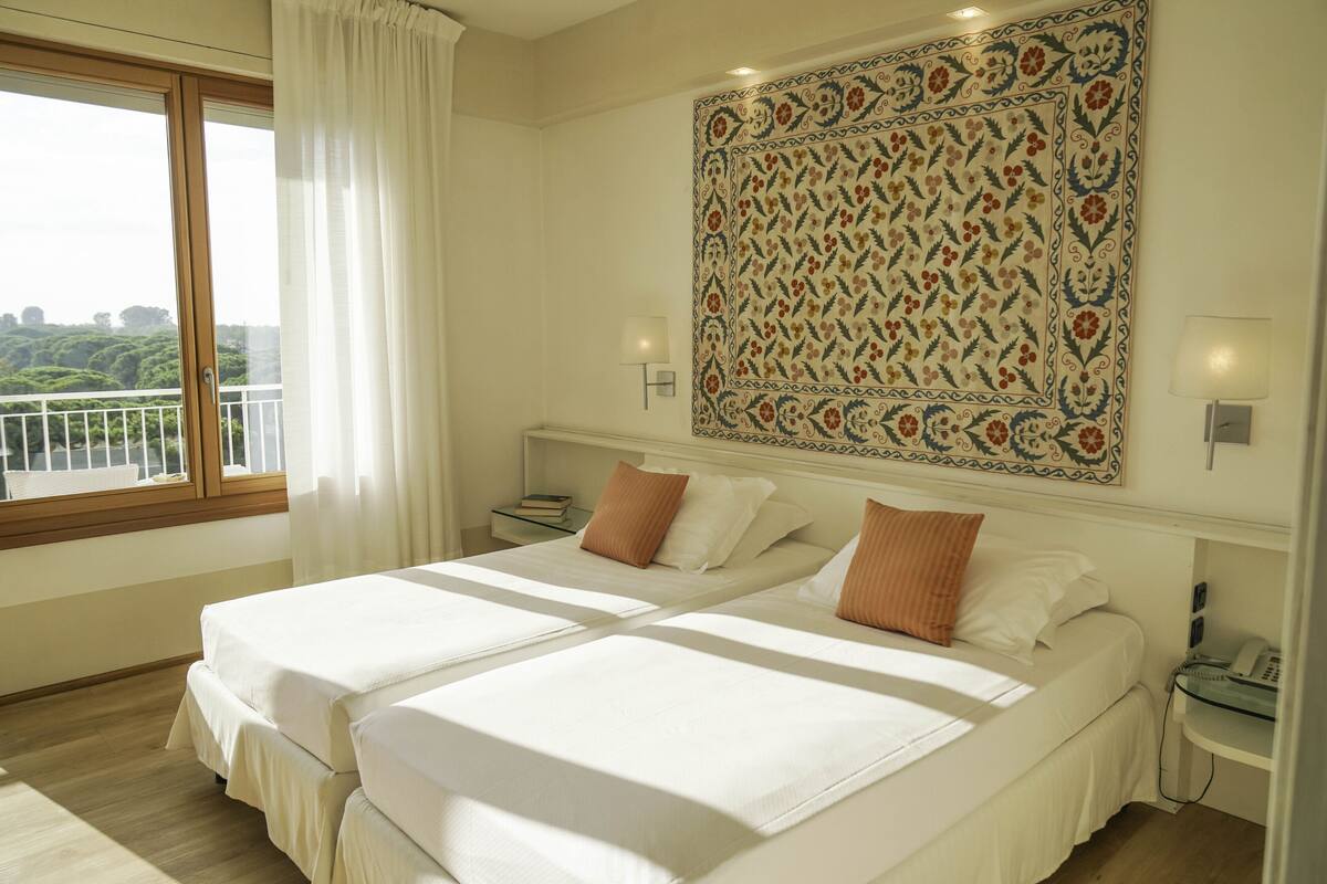 A hotel room with two beds, a large window with curtains, a patterned tapestry on the wall, and a small bedside table with a lamp and a phone.