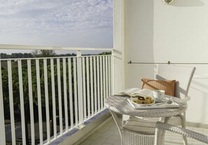 A balcony with a white railing, a wicker chair, a small table with a book, a cup, and a plate with food. - Union Lido Art & Park Hotel (Cavallino-Treporti)