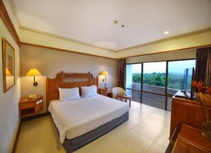 A hotel room with a large bed, two bedside tables with lamps, a small round table, and a view of a forest through large windows.