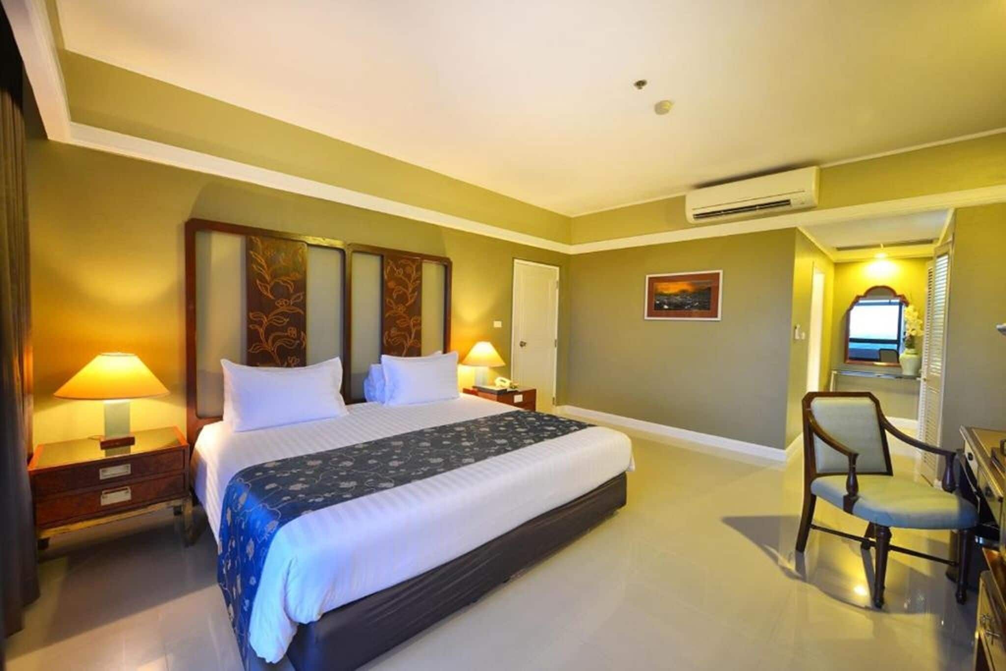 A hotel room with a large bed, two bedside tables with lamps, a chair, a desk, and an air conditioning unit.