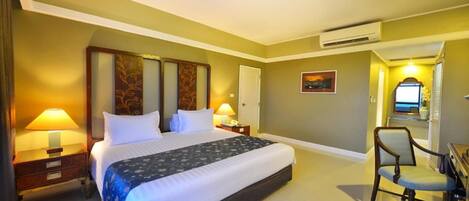 A hotel room with a large bed, two bedside tables with lamps, a chair, a desk, and an air conditioning unit.