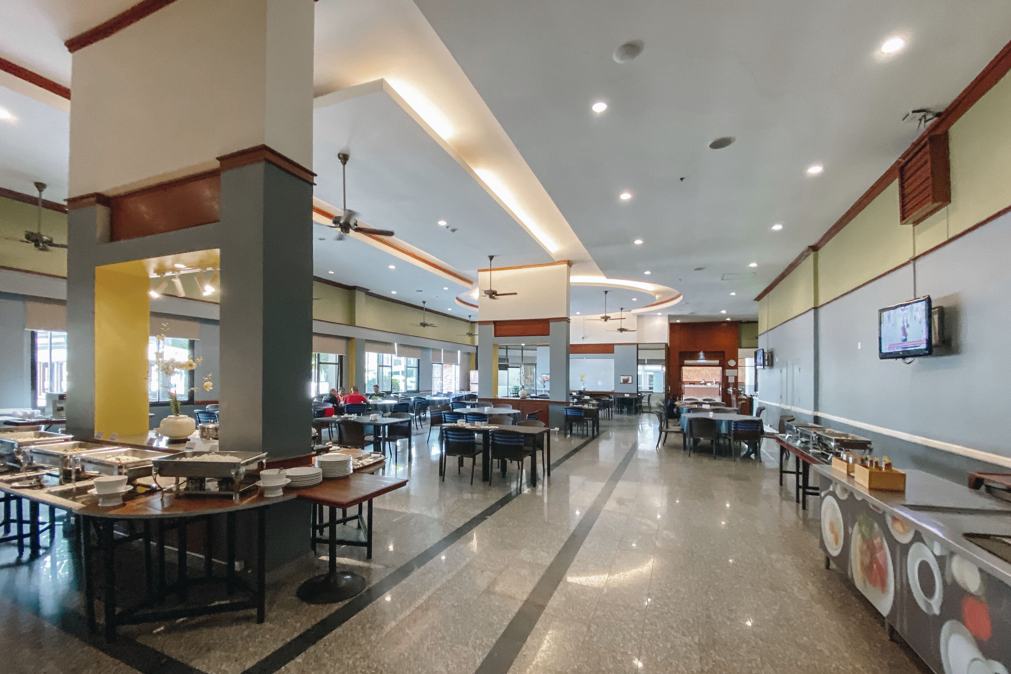 Una cafetería amplia con varias mesas y sillas, ventiladores de techo y una pantalla de televisión en la pared.