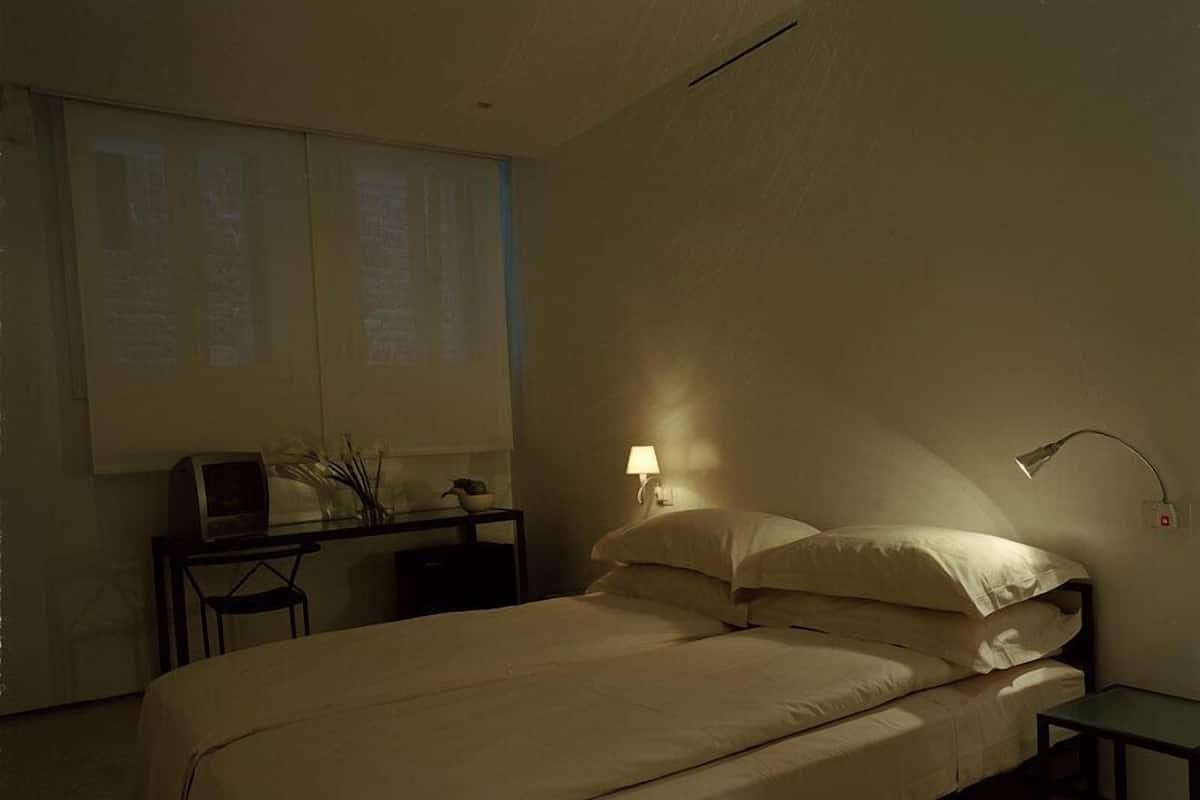 Un dormitorio con una cama, un escritorio con computadora, una lámpara y una ventana con persianas.