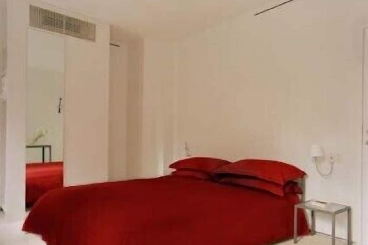 Un dormitorio moderno con una colcha roja, una mesita de noche pequeña y una pared blanca.