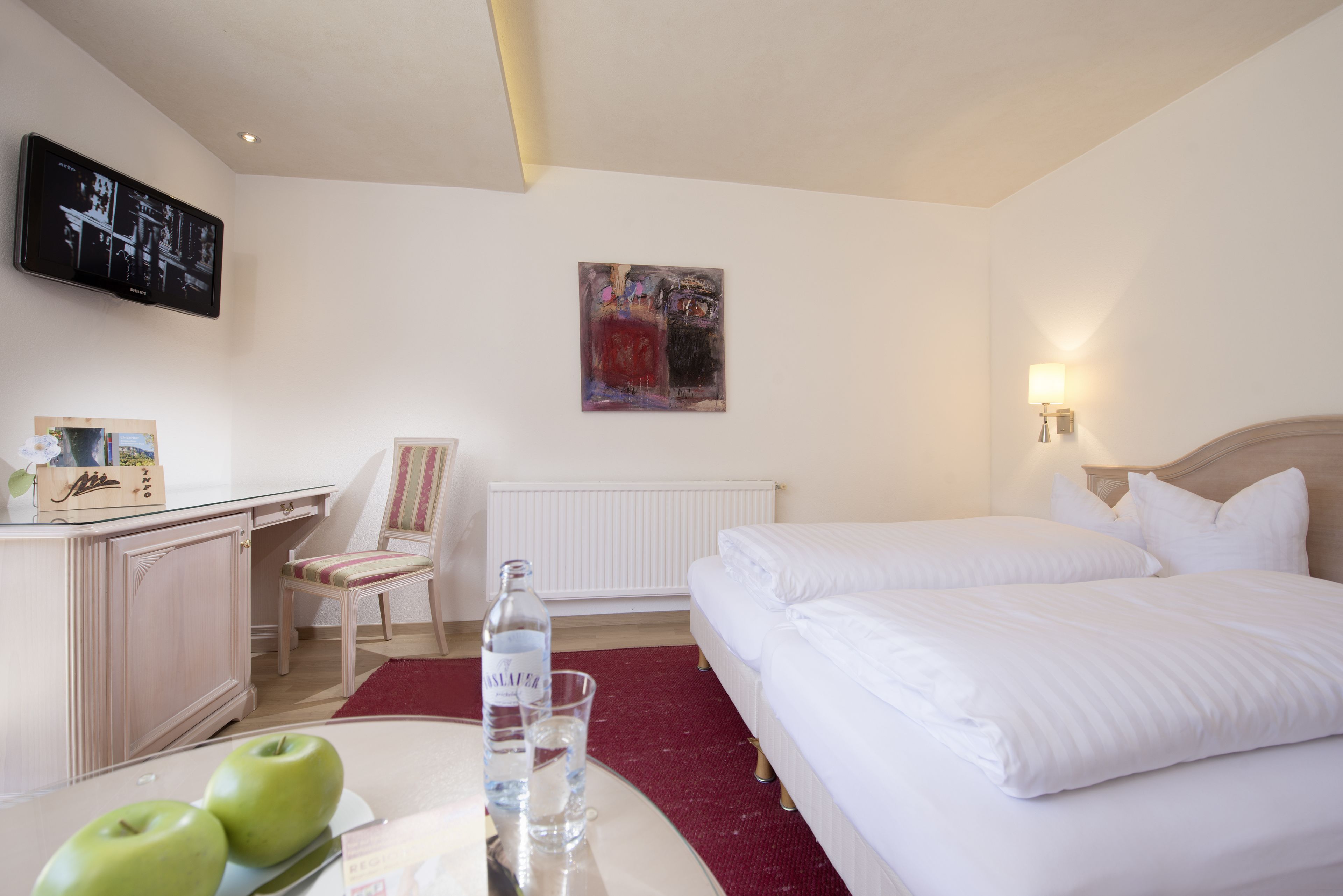 A hotel room with a bed, a desk with a chair, a television mounted on the wall, and a painting above the bed.