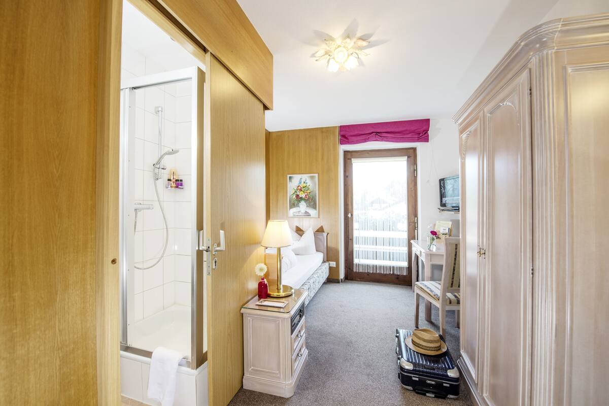 A hotel bathroom with a shower, a vanity with a mirror, a bed, and a suitcase on the floor.