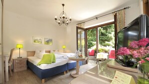 A hotel room with a bed, bedside lamps, a desk, a chair, a window with a view of greenery, and a chandelier. - Hotel Maximilian (Ehenbichl)