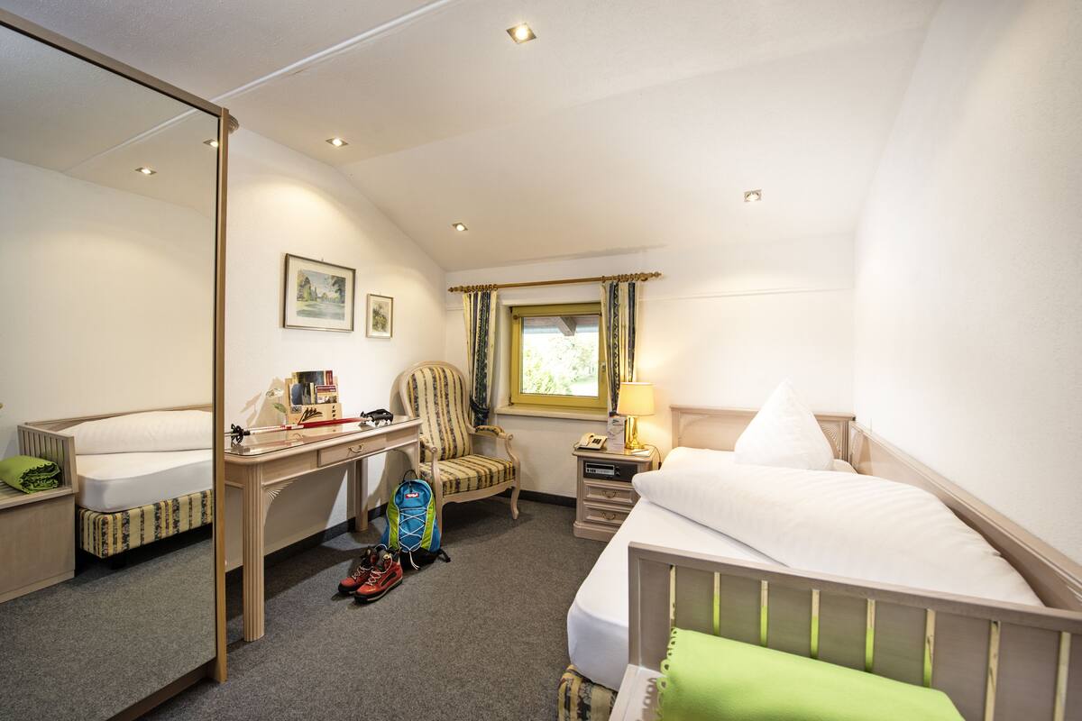 A hotel room with a bed, a desk, a chair, a small table with a lamp, a window with curtains, and a mirror.