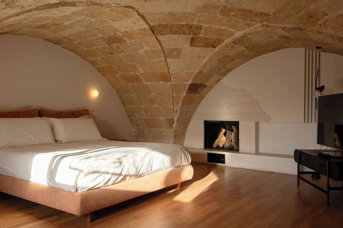 A bedroom with a stone vaulted ceiling, a bed with white linens, a wooden desk, and a fireplace.