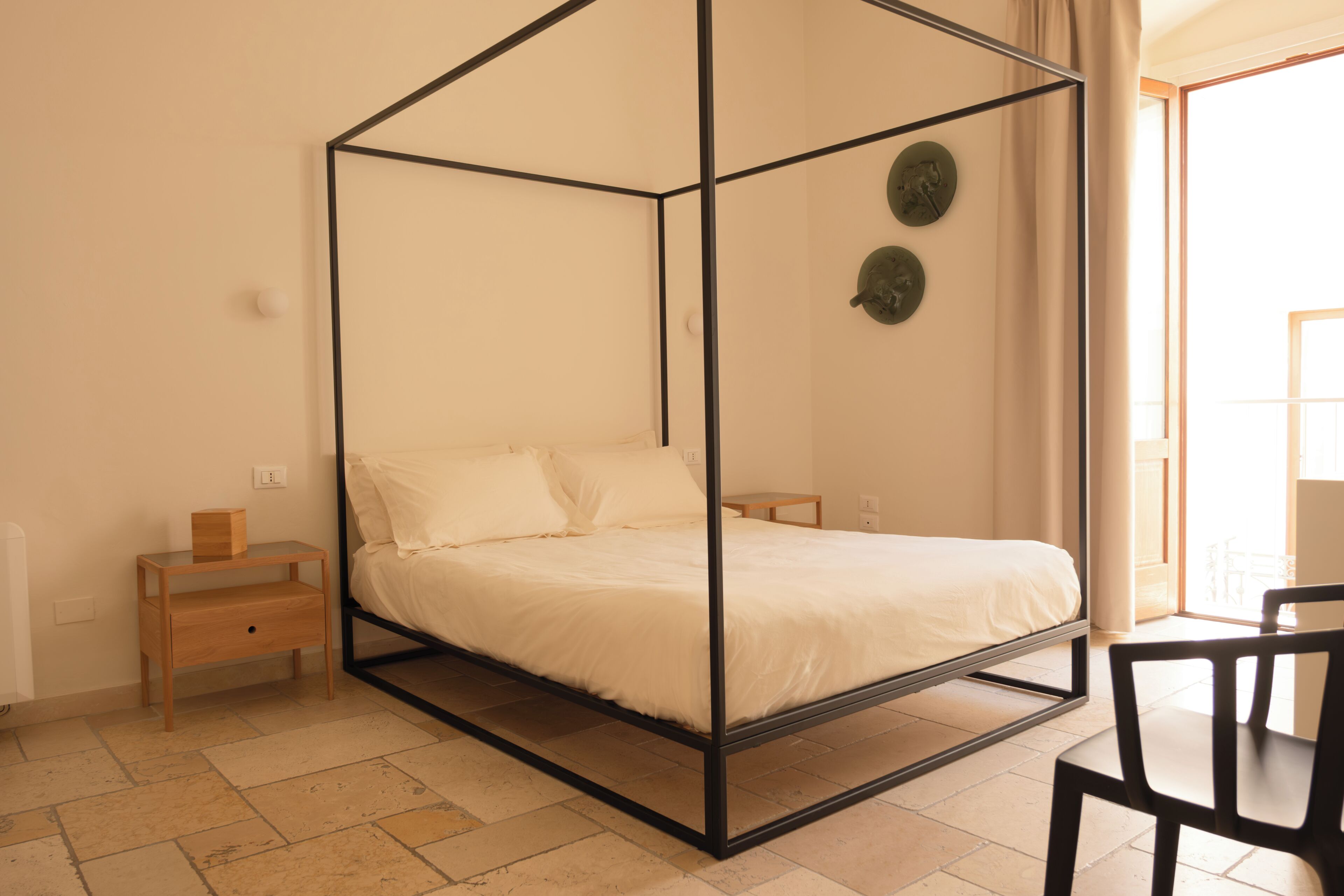 Een kamer met een hemelbed, een houten nachtkastje, een stoel en uitzicht op buiten.