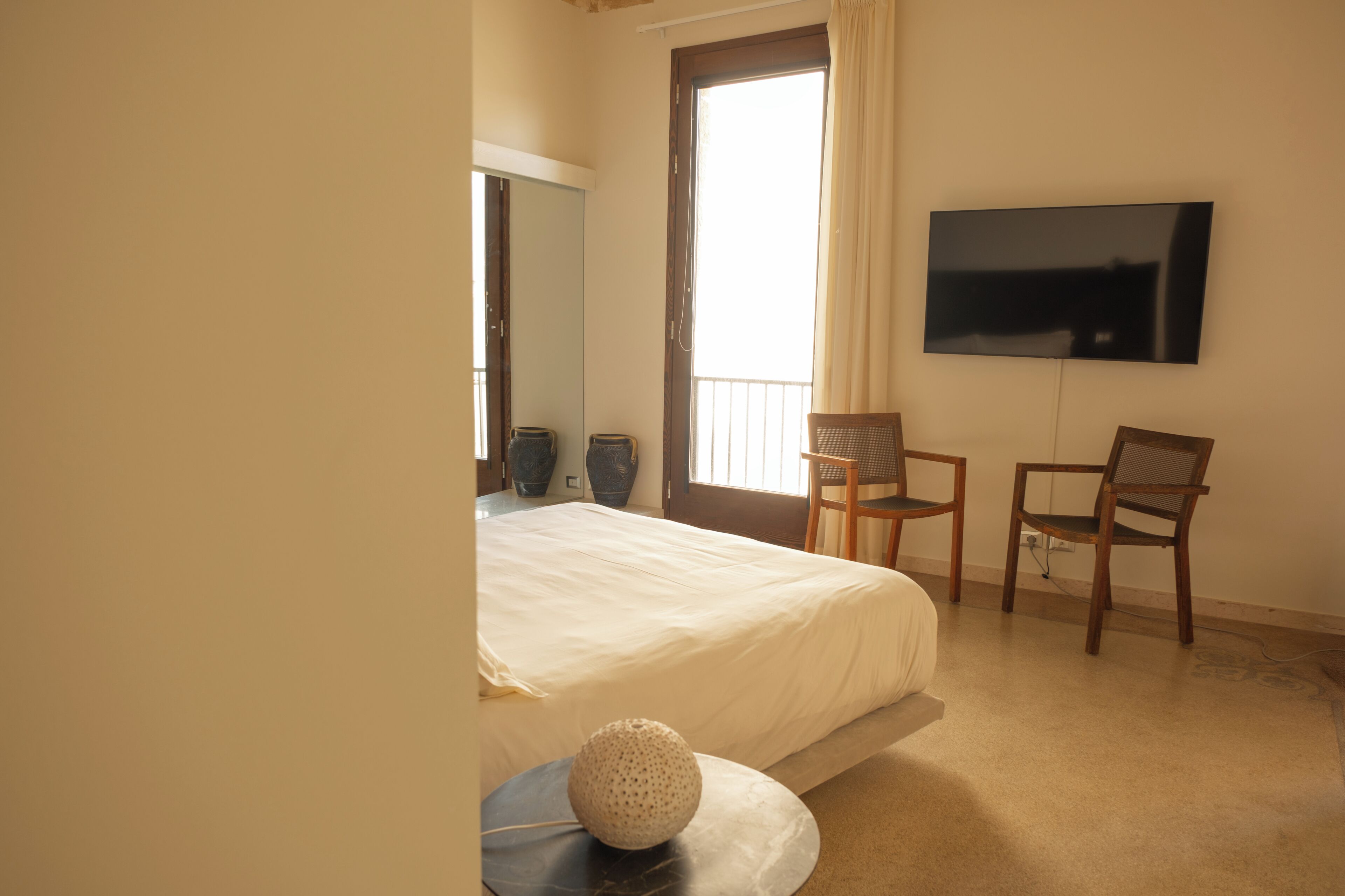 A bedroom with a bed, two chairs, a TV on the wall, and a large window.