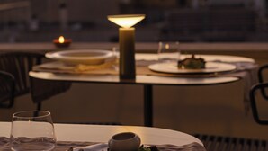 Mesa dispuesta para la cena, con una vela y una lámpara, con vistas a un paisaje urbano al atardecer.