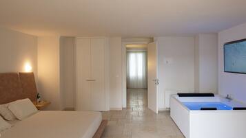 Habitación de hotel con cama, mesita de noche, ventana grande, baño con bañera y pasillo que conduce a otra habitación.