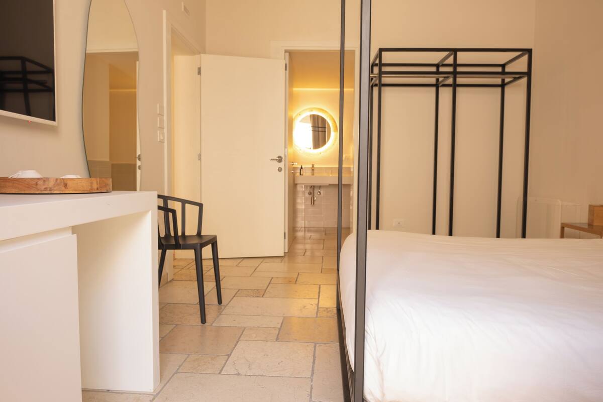 A hotel room with a bed, a desk with a chair, a mirror, and a bathroom with a round mirror.