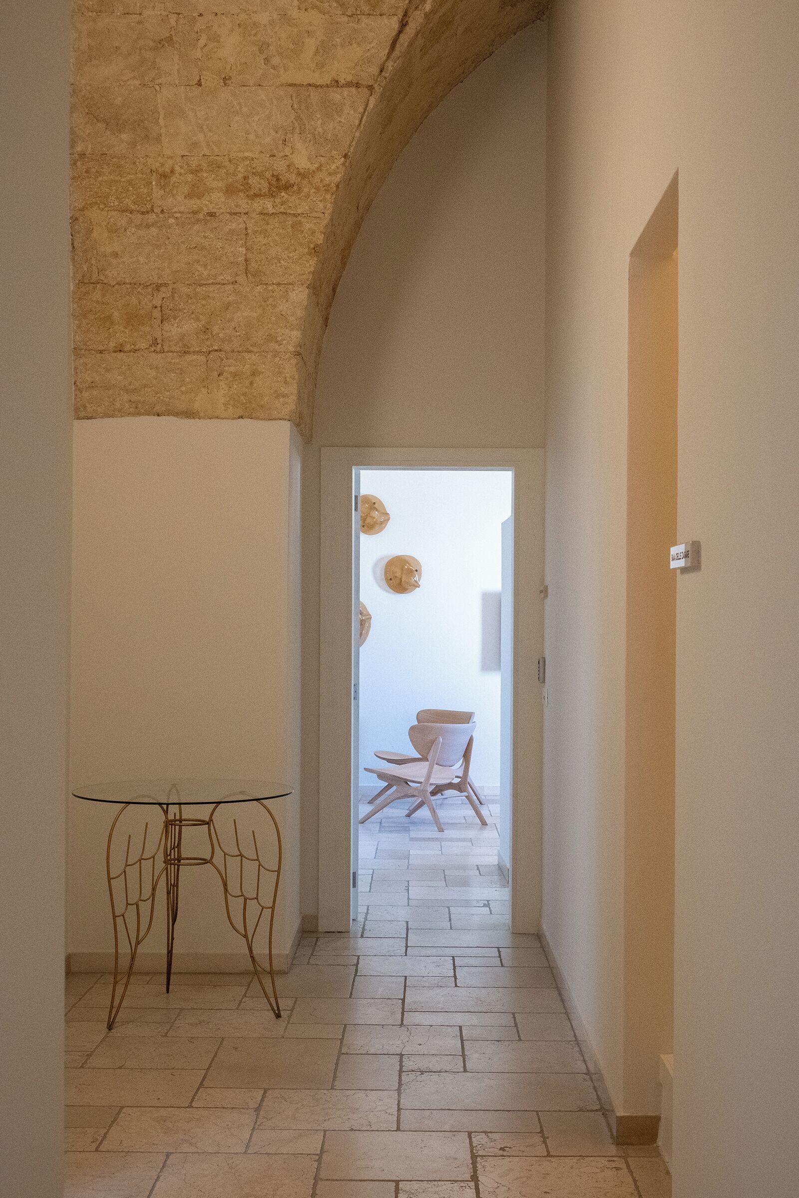 A hallway with a stone wall, a glass table with metal legs, and a door with a sign.