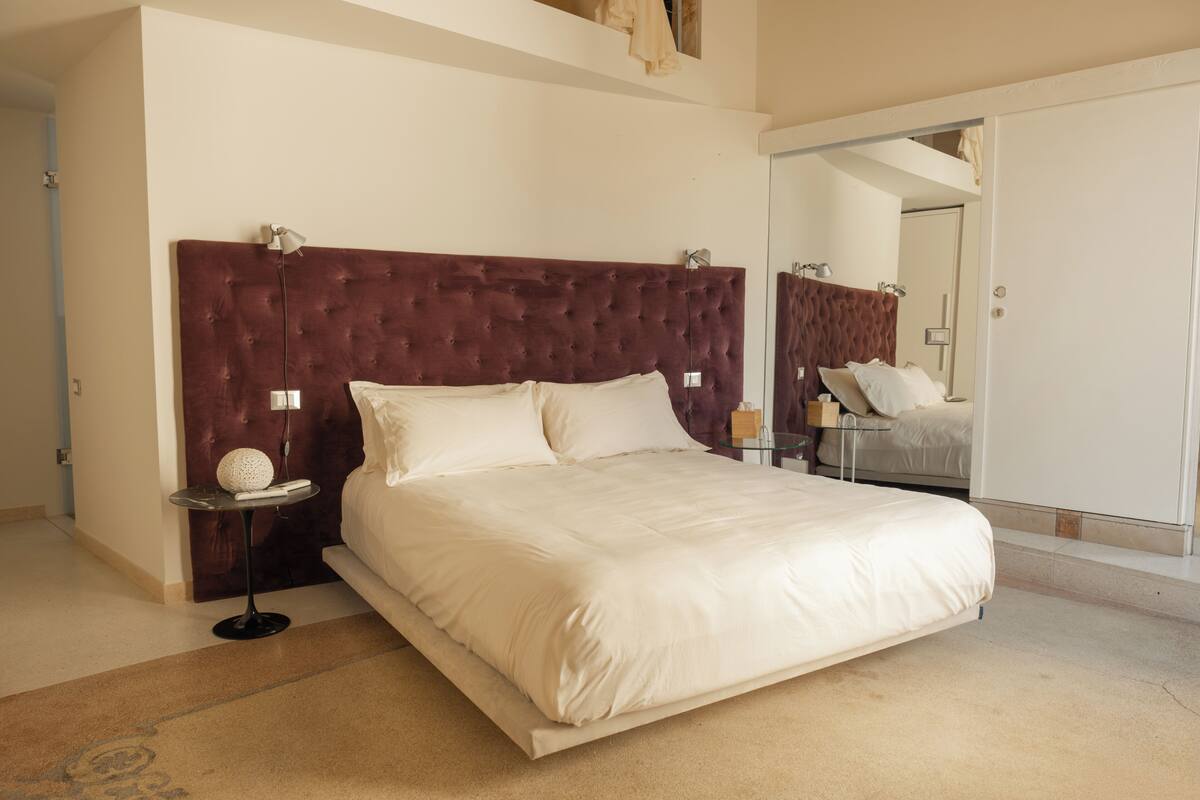 A bedroom with a large bed, a bedside table with a lamp, a mirror, and a wardrobe.