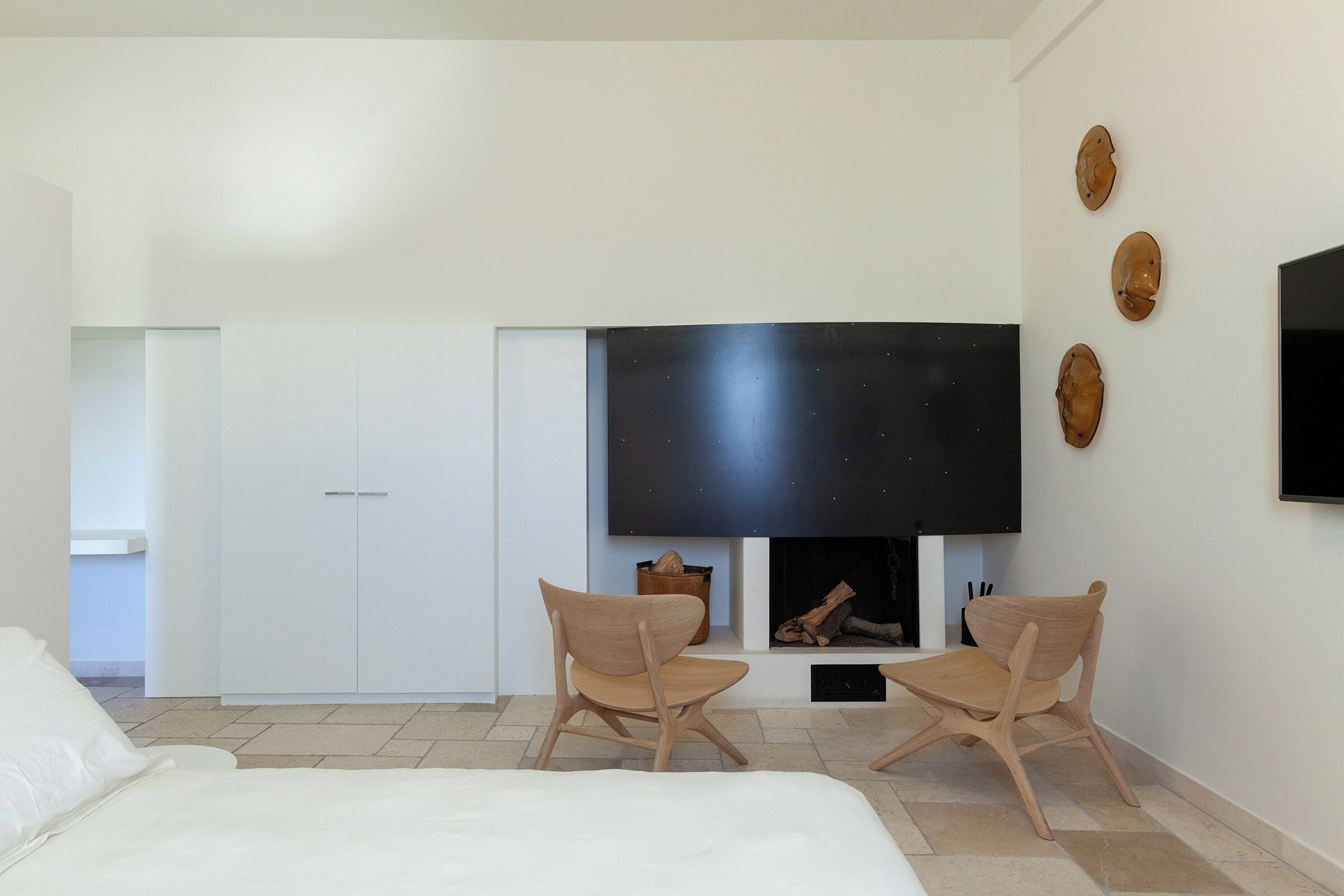A modern bedroom with a bed, two wooden chairs, a TV, and a fireplace.