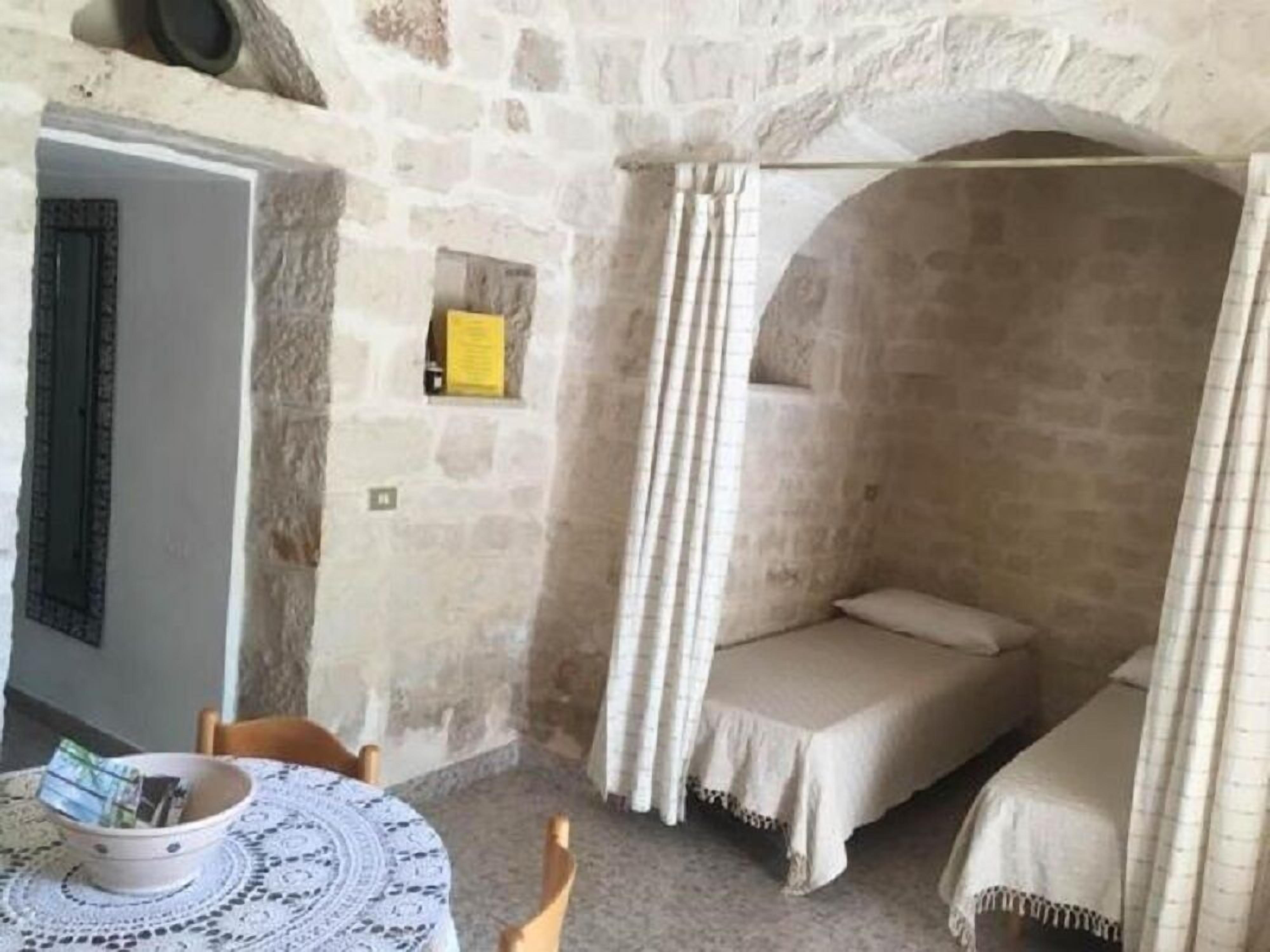 Una habitación rústica con pared de piedra, una pequeña mesa con un cuenco, una cama con dosel y una silla.