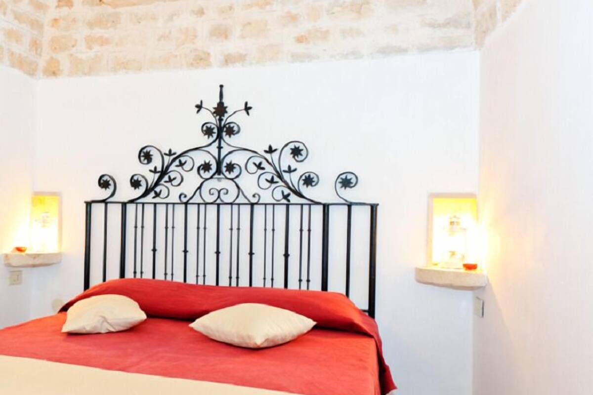 A bed with a red and beige bedspread, two pillows, and a decorative metal headboard. There are two wall-mounted nightstands with lamps.