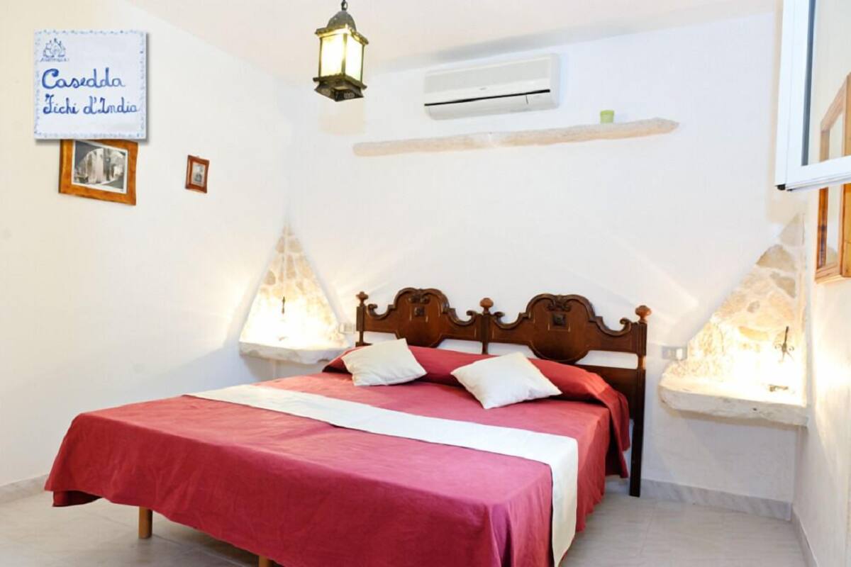 A single bed with a wooden headboard, a red bedspread, and two bedside lamps. There is a small shelf with a green object on it, and a wall-mounted air conditioning unit.
