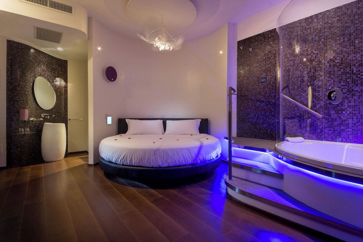 A modern bedroom with a circular bed, a large mirror, a glass-enclosed shower, and a small bathroom with a sink and mirror.