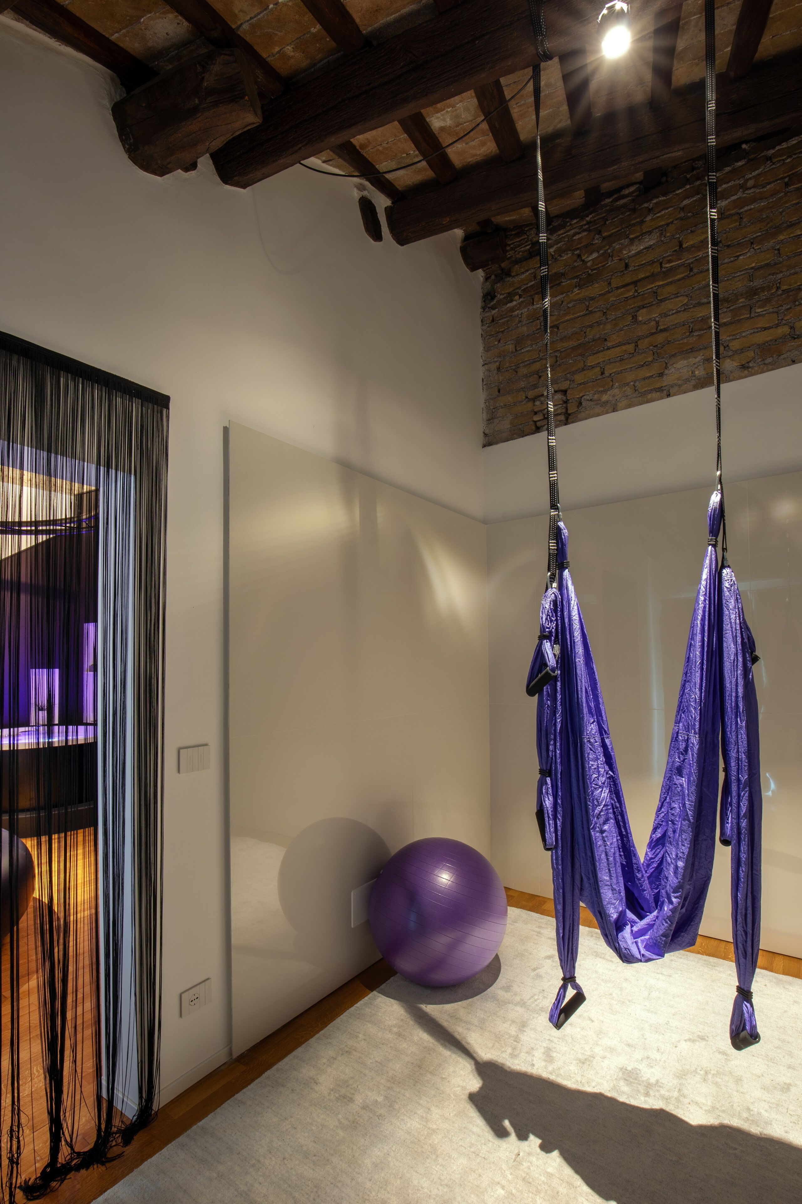 Una habitación con una hamaca de tela colgante para hacer ejercicio, una pelota grande de gimnasia y una cortina con líneas verticales.