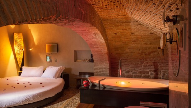 A cozy room with a brick ceiling, a bed with white linens, a bedside table, and a modern bathroom with a large mirror and sink.
