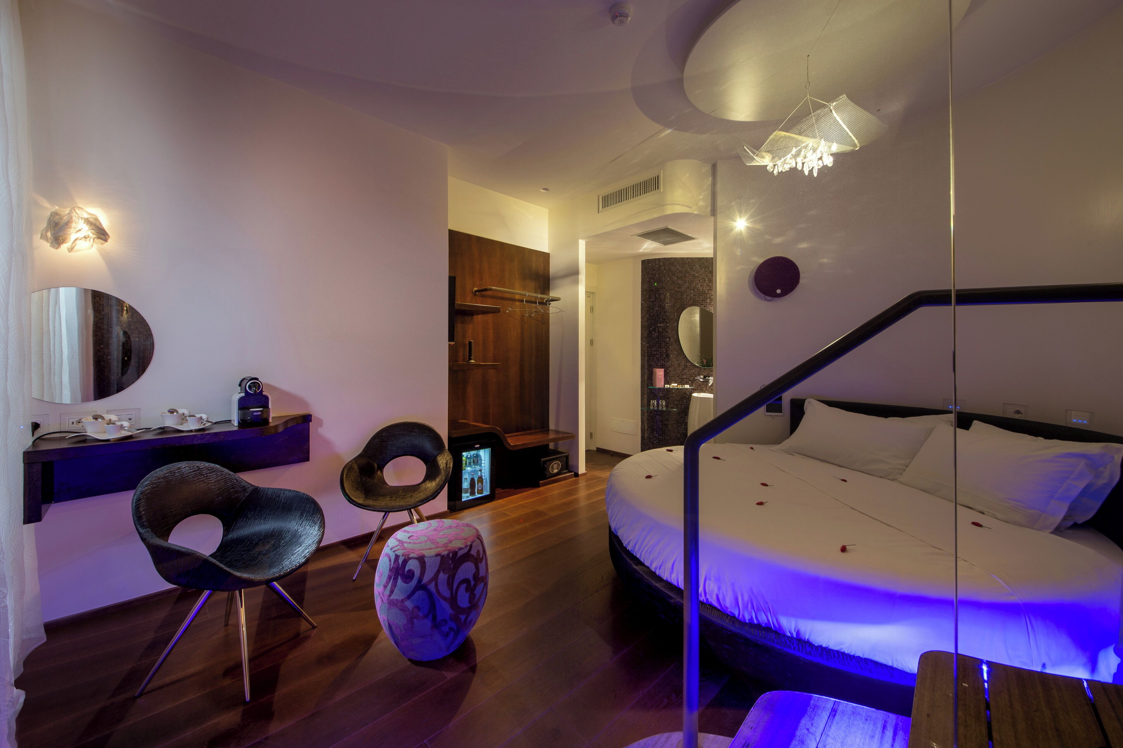 Une chambre à coucher moderne avec un grand lit, un bureau avec une chaise, un pouf rond et un miroir.