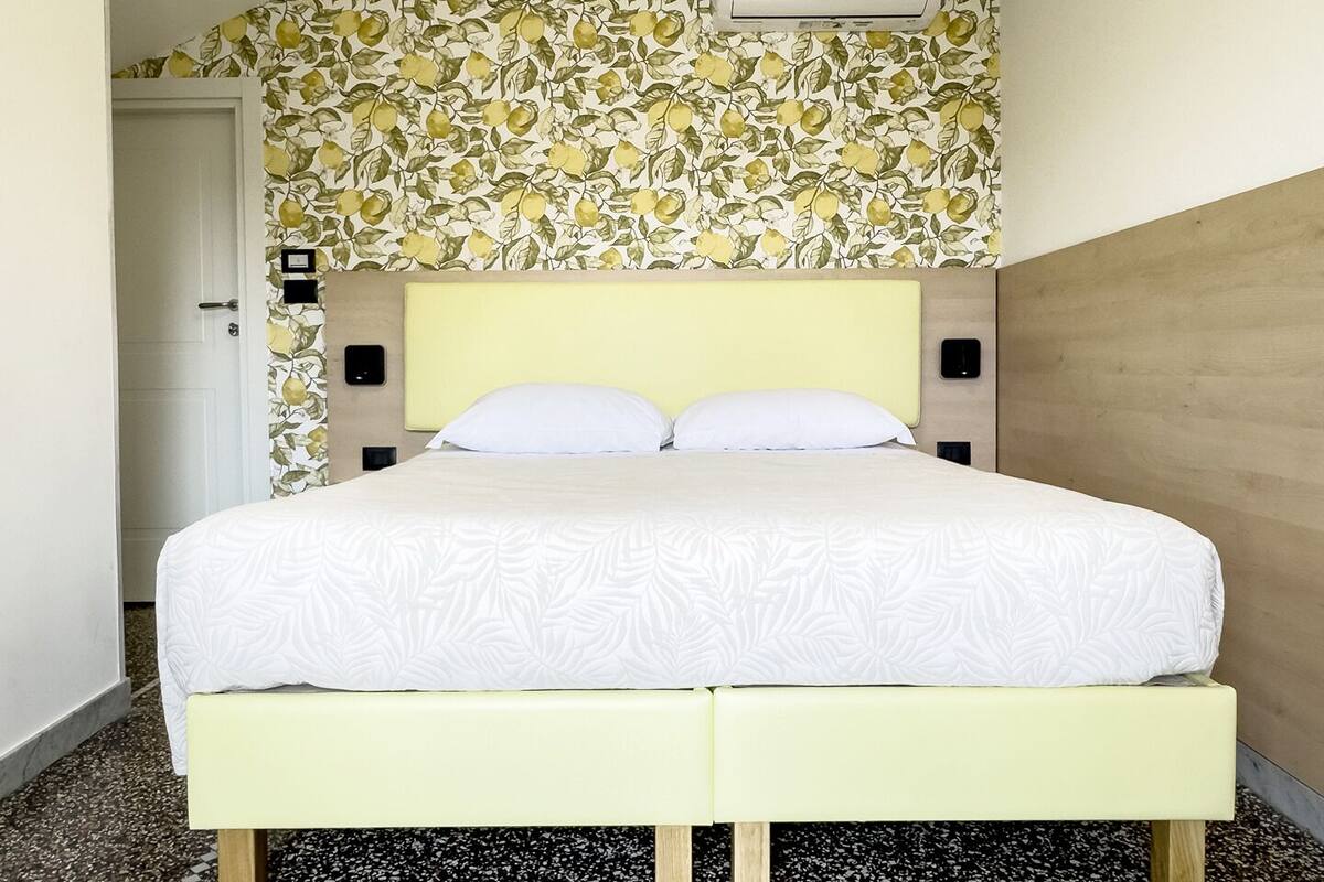 A bed with a white bedspread, two white pillows, a yellow headboard, and a floral wallpaper pattern above.