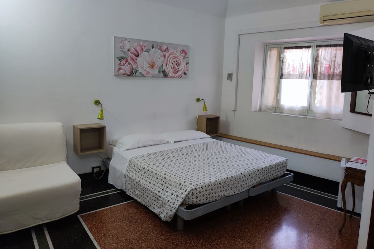 A bedroom with a bed, a chair, a painting, a window with curtains, and a television.