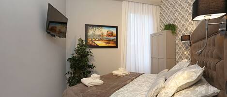 A neatly arranged bedroom with a bed, a television mounted on the wall, a painting, and a plant.