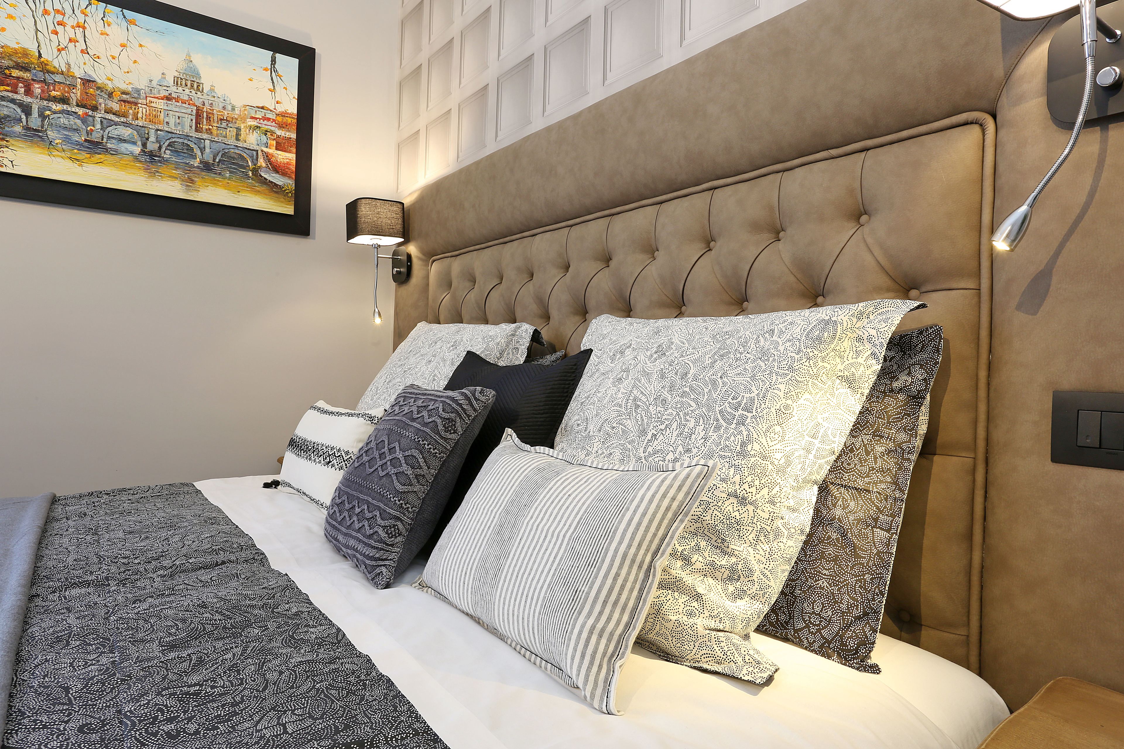 A bedroom with a tufted headboard, patterned bedding, and a cityscape painting on the wall.