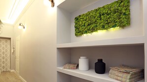 A hallway with a white wall, a green moss wall art, and a shelf with a white vase and a black vase. - Capricci Romani (Rome)