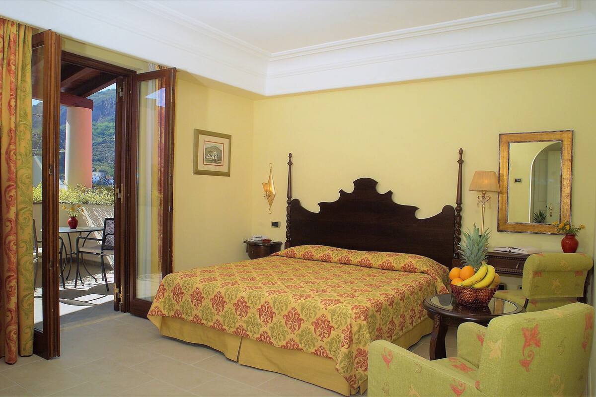 A hotel room with a bed, a chair, a table with fruit, and a balcony visible through an open door.