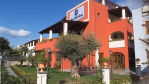 A red building with a sign that reads "Hotel de la Mer." It has a balcony with plants and a staircase leading to the entrance. - Grand Hotel Arciduca (Lipari)