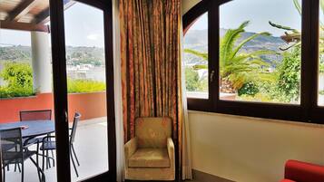 A room with a view of a mountainous landscape through large windows, a chair, a table with chairs, and a red sofa.