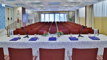 A conference room with rows of red chairs, a white table with a patterned tablecloth, microphones, and decorative plants.