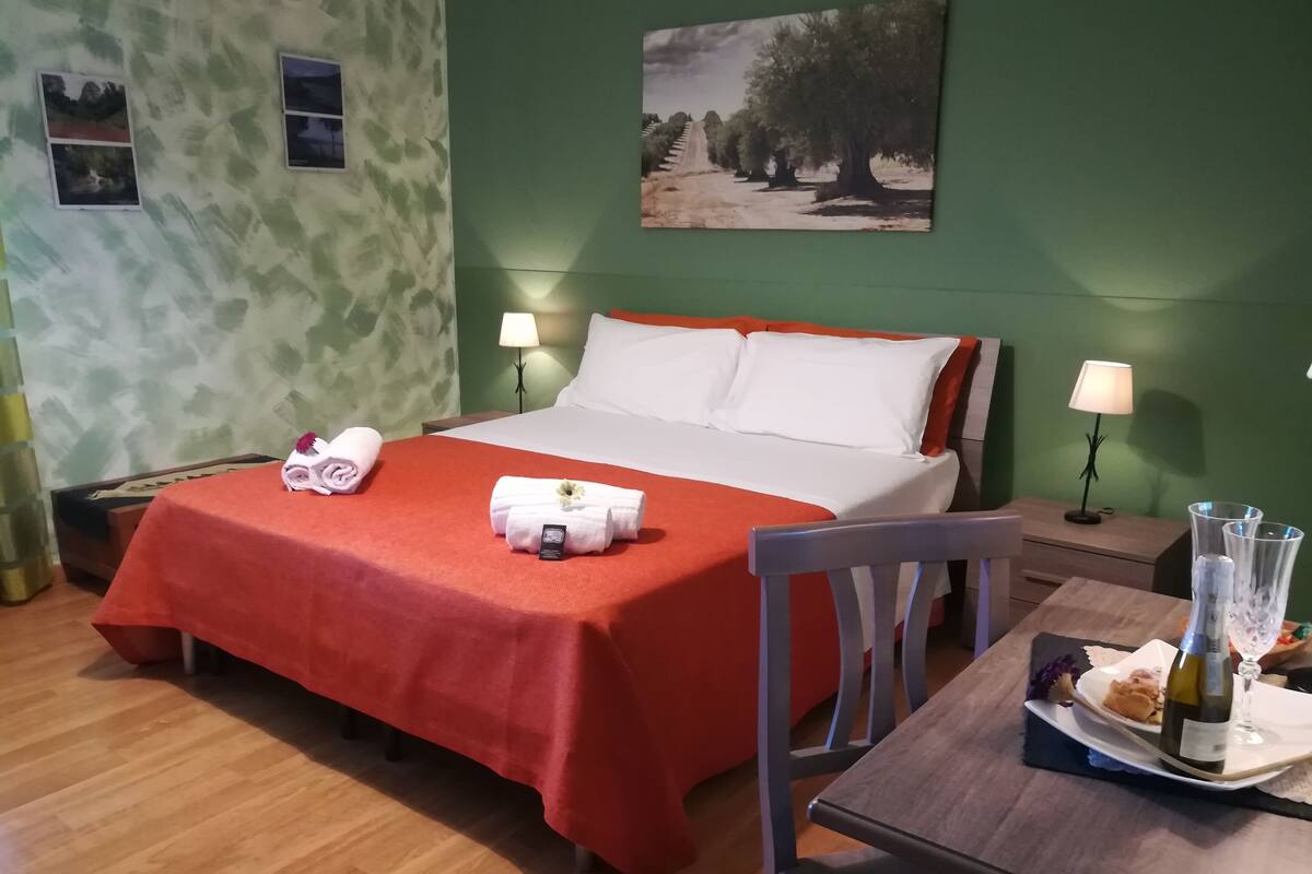 A hotel room with a bed, two bedside lamps, a wooden nightstand, a dining table with a bottle and glass, and a chair.