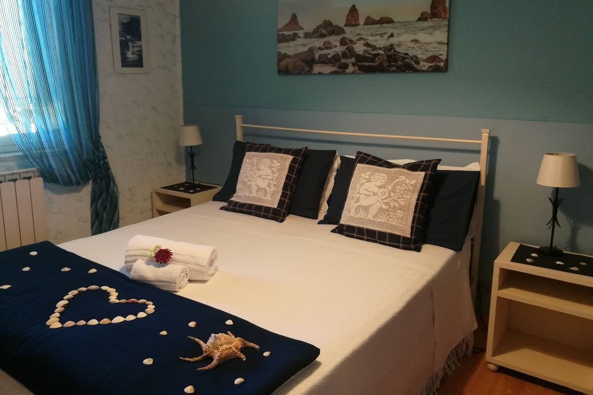 A neatly made bed with a heart-shaped arrangement of stones and a seashell, a painting of a coastal scene, and a window with blue curtains.