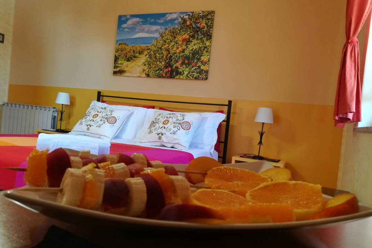 A hotel room with a bed, pillows, a bedside table with a lamp, and a fruit platter on the bed.