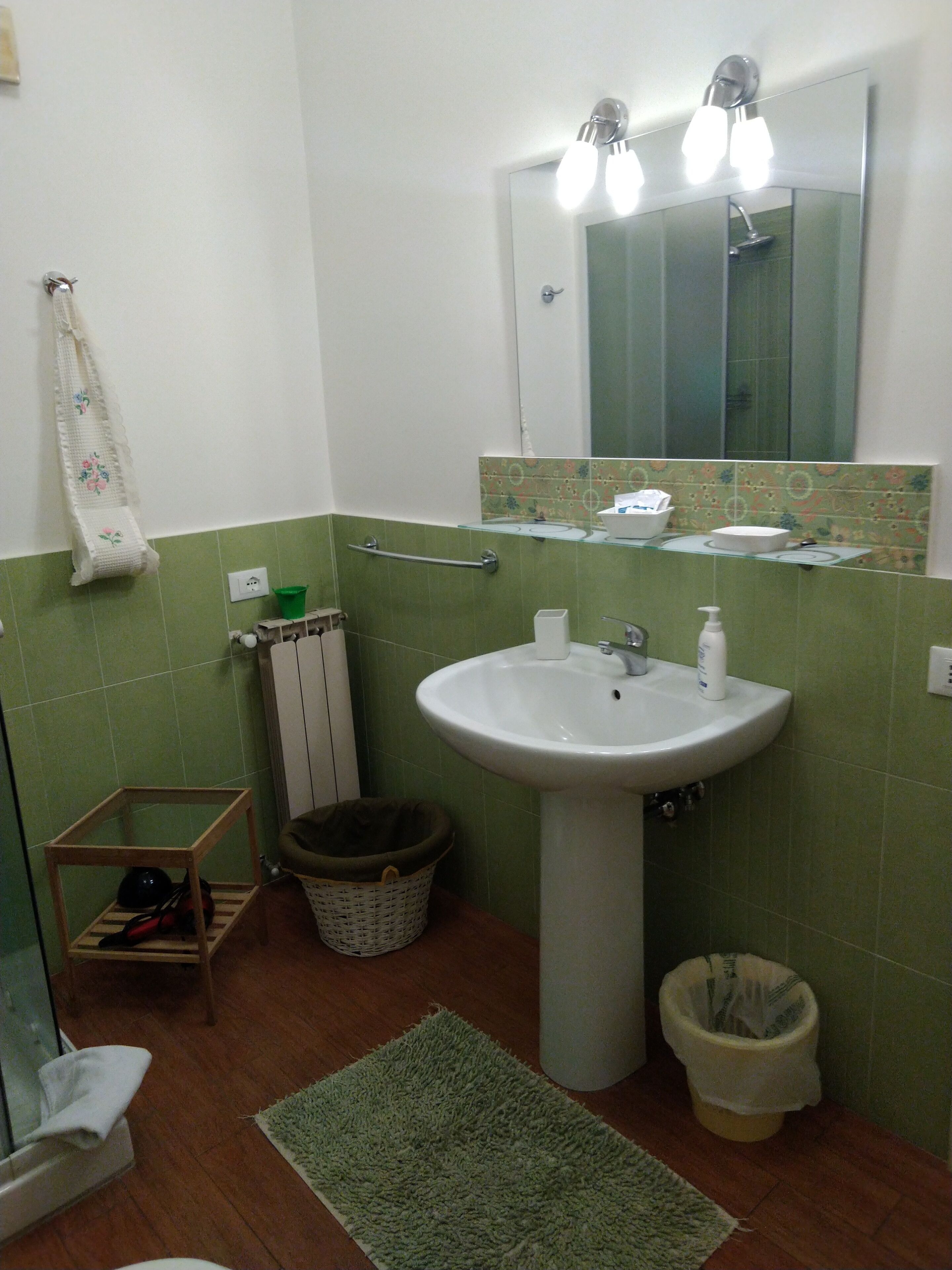 Un baño con lavamanos, espejo, azulejos verdes, toallero, estante pequeño y cubo de basura.