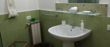 Un baño con lavamanos, espejo, azulejos verdes, toallero, estante pequeño y cubo de basura.
