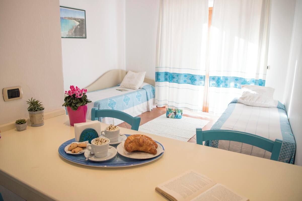 A hotel room with two beds, a dining area with breakfast, a small potted plant, and a window with curtains.